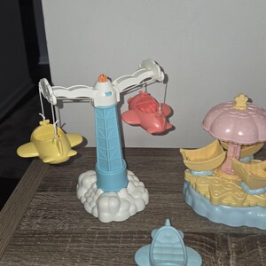 Puede incluir: Un conjunto de aviones de juguete coloridos y una estructura similar a un carrusel. El carrusel tiene una torre azul con una parte superior blanca y nubes en la base, con aviones amarillos y rojos colgando de &eacute;l. Un avi&oacute;n azul est&aacute; en la superficie.