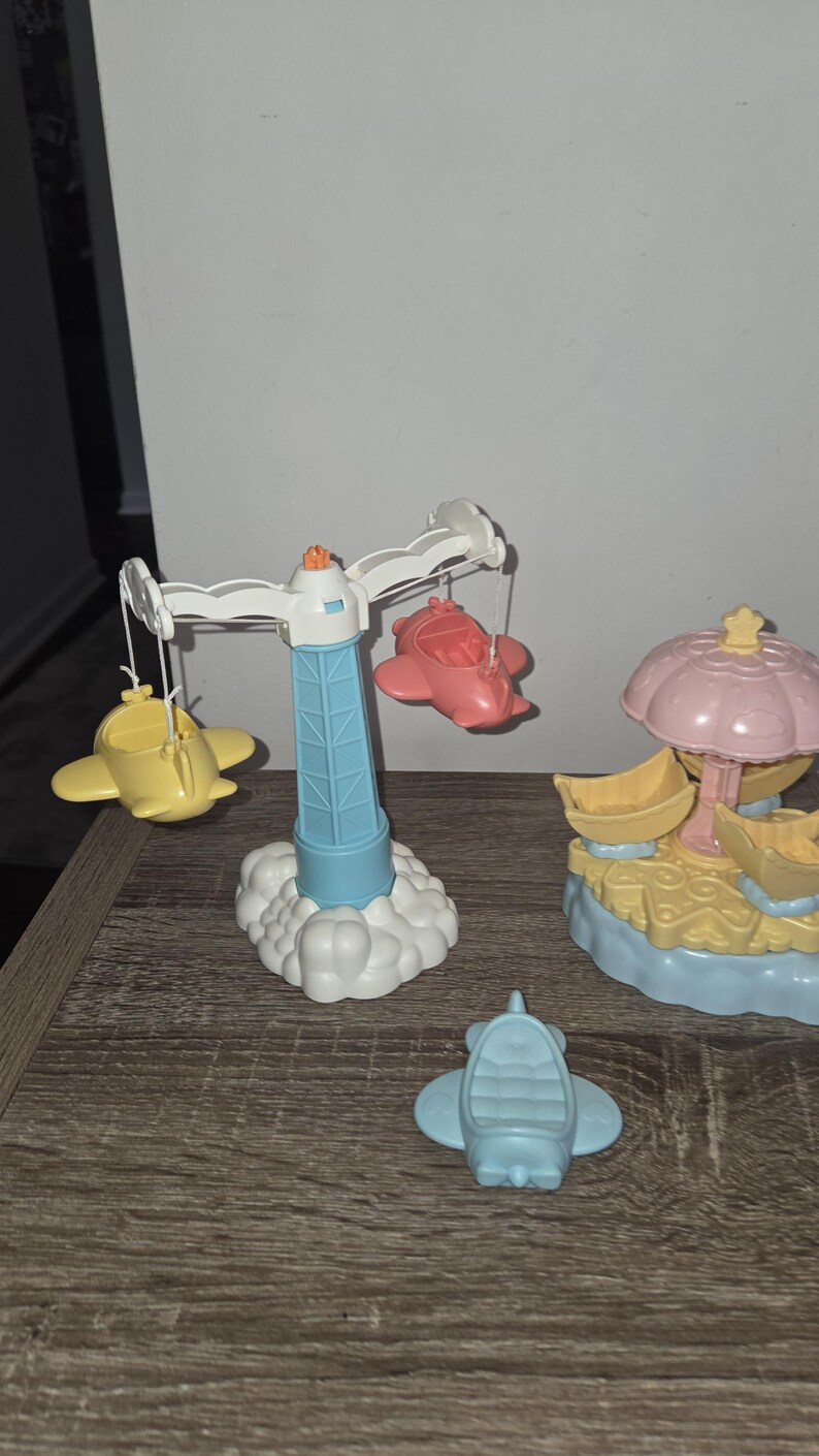 Puede incluir: Un conjunto de aviones de juguete coloridos y una estructura similar a un carrusel. El carrusel tiene una torre azul con una parte superior blanca y nubes en la base, con aviones amarillos y rojos colgando de &eacute;l. Un avi&oacute;n azul est&aacute; en la superficie.