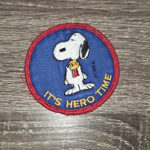 May include: Round patch featuring Snoopy in a hero pose, with the text "IT'S HERO TIME". The patch has a blue background with a red border and Snoopy in white and black.
