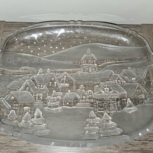 May include: A clear glass serving tray with a winter village scene etched into the surface. The tray features a detailed depiction of houses, trees, and a snowy landscape, with handles on either side. The tray is oval-shaped.