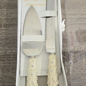 May include: A two-piece cake serving set featuring a serrated knife and a triangular server with stainless steel blades. The handles are cream-colored with a textured floral and ribbon design, accented by small embedded clear crystals.