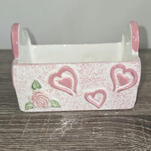 May include: A rectangular ceramic basket with a white interior and a pink speckled exterior. The front features three raised pink heart outlines and a single pink rose with green leaves. The basket has two upright handles with pink accents.