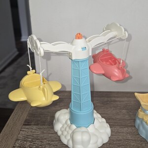 Puede incluir: Un carrusel de juguete colorido con una torre azul claro y una base de nube blanca. El carrusel tiene brazos blancos con aviones amarillos y rosas adjuntos. La parte superior de la torre es naranja.