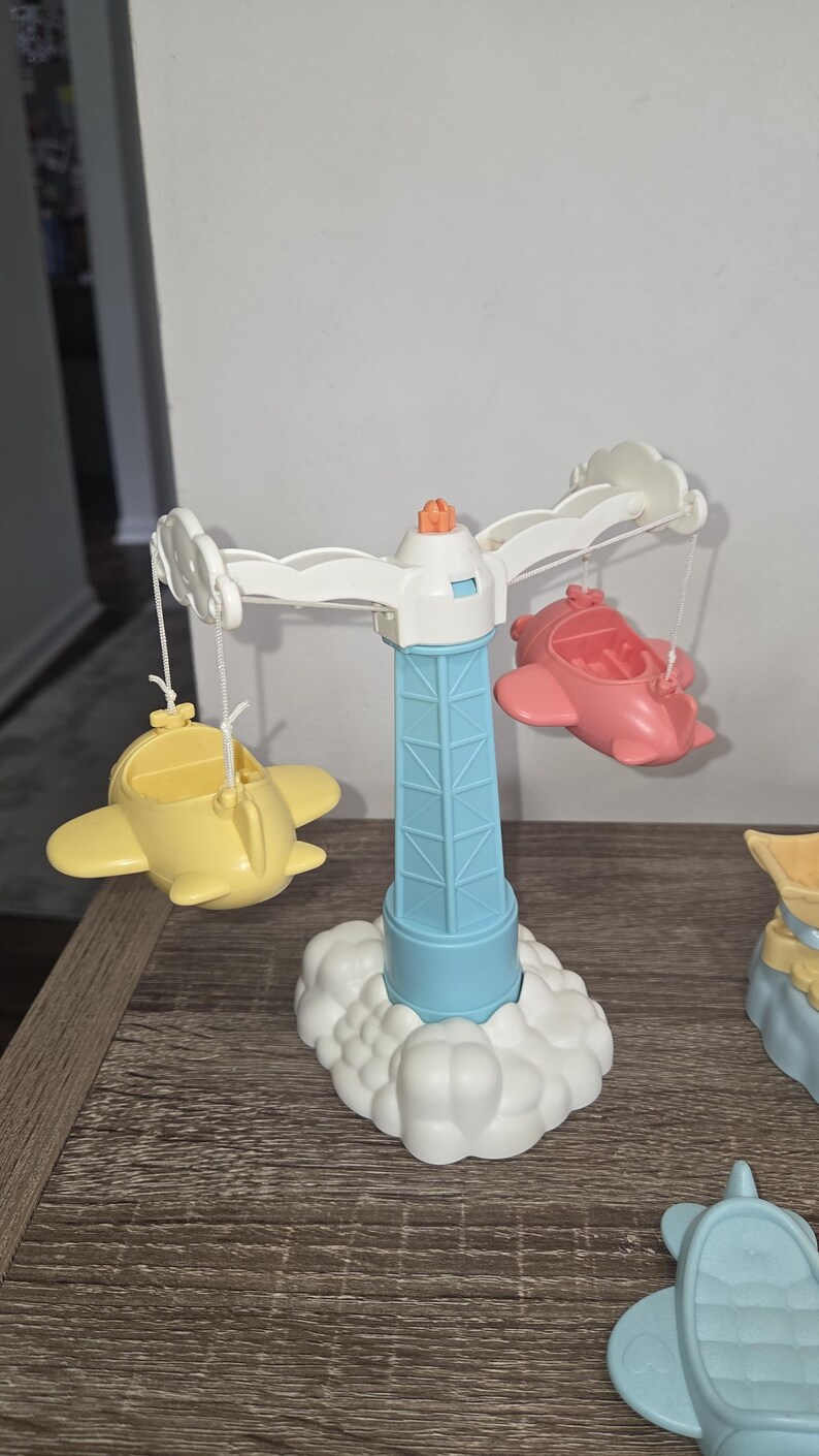 Puede incluir: Un carrusel de juguete colorido con una torre azul claro y una base de nube blanca. El carrusel tiene brazos blancos con aviones amarillos y rosas adjuntos. La parte superior de la torre es naranja.