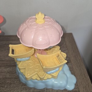 Puede incluir: Un juguete de carrusel de colores pastel con una parte superior rosa en forma de paraguas y asientos amarillos. La base es azul claro y amarilla, con un acento en forma de estrella en la parte superior. El juguete es de pl&aacute;stico.