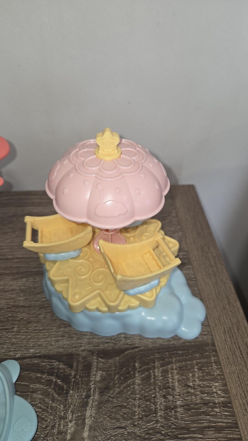 Puede incluir: Un juguete de carrusel de colores pastel con una parte superior rosa en forma de paraguas y asientos amarillos. La base es azul claro y amarilla, con un acento en forma de estrella en la parte superior. El juguete es de pl&aacute;stico.