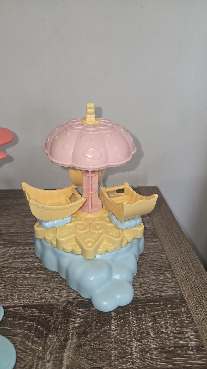 Puede incluir: Un juguete de carrusel en colores pastel con una parte superior rosa en forma de paraguas y dos asientos amarillos en forma de barco. La base es azul claro y amarilla, con detalles de nubes y remolinos. Un juguete infantil.