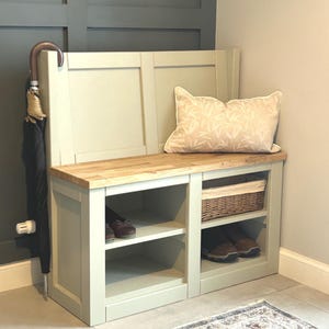 May include: A light green wooden bench with a natural wood top, ideal for an entryway. It features open storage with shelves, a woven basket, and a decorative pillow. An umbrella is positioned next to the bench. The bench is approximately 120 cm wide.