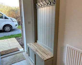 May include: A tall, sage green hallway storage unit featuring a vertical paneled backboard with a row of black metal coat hooks. The base consists of a bench with a light-toned wooden seat and enclosed storage compartments underneath.