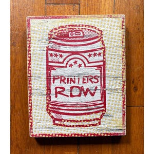 May include: A rectangular wooden sign with a distressed finish. The sign features a red and white illustration of a soda can with the words "PRINTERS ROW" in a bold font. The background is a cream color with gold dots.