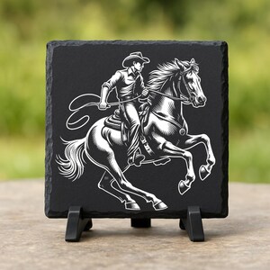 May include: Black square slate coaster featuring a white illustration of a cowboy riding a horse. The cowboy is wearing a hat and holding a lasso. The horse is rearing up on its hind legs. The coaster is displayed on a black stand.