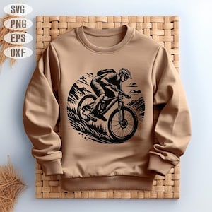Op de afbeelding: Een bruin sweatshirt met een zwarte afbeelding van een persoon die met een mountainbike over een pad rijdt. De afbeelding is in een cirkel met een bergketen op de achtergrond.
