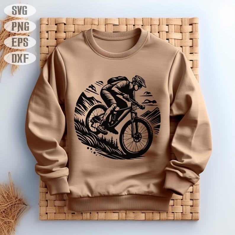 Op de afbeelding: Een bruin sweatshirt met een zwarte afbeelding van een persoon die met een mountainbike over een pad rijdt. De afbeelding is in een cirkel met een bergketen op de achtergrond.