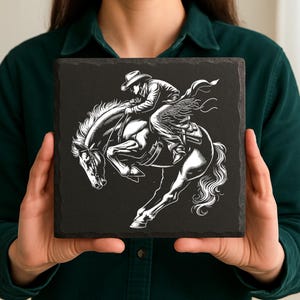 May include: Square black slate coaster featuring a detailed black and white illustration of a cowboy riding a bucking horse. The cowboy is wearing a hat and chaps. The horse is rearing up, with its front legs in the air.