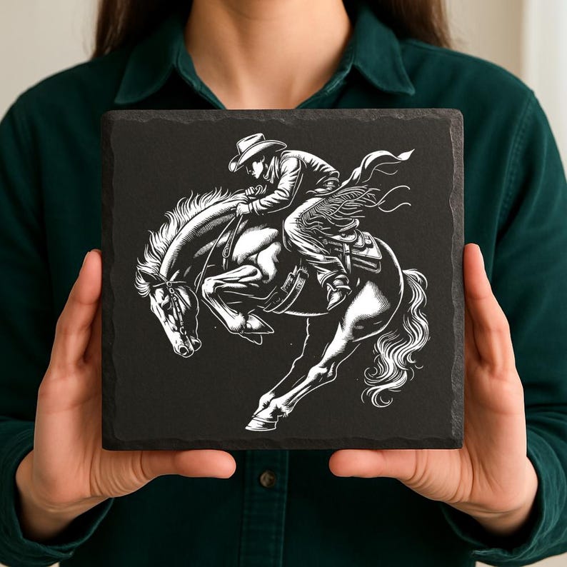 May include: Square black slate coaster featuring a detailed black and white illustration of a cowboy riding a bucking horse. The cowboy is wearing a hat and chaps. The horse is rearing up, with its front legs in the air.