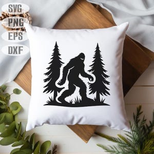 May include: White square pillow featuring a black silhouette of Bigfoot walking between two pine trees. The design is a graphic illustration. The pillow is on a wooden surface with greenery in the background. The top left corner has the text "SVG, PNG, EPS, DXF".