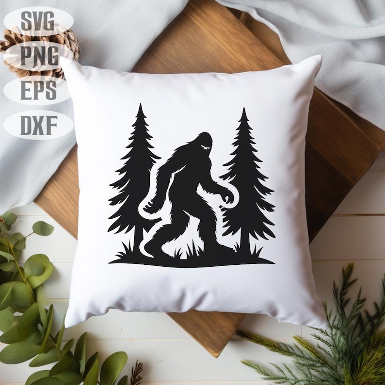 May include: White square pillow featuring a black silhouette of Bigfoot walking between two pine trees. The design is a graphic illustration. The pillow is on a wooden surface with greenery in the background. The top left corner has the text "SVG, PNG, EPS, DXF".