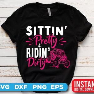May include: Black t-shirt with the text "Sittin' Pretty & Ridin' Dirty" in pink lettering. A pink and black side-by-side vehicle is pictured below the text.