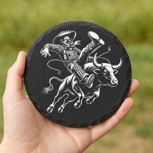 May include: A black round coaster with a white illustration of a rodeo clown riding a bull. The clown wears a sombrero and striped pants. The bull is bucking. The coaster is held in a hand.