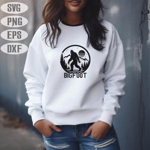 May include: White sweatshirt with a black Bigfoot graphic. The graphic features a silhouette of Bigfoot in a forest scene with a sun and the word "BIGFOOT". The sweatshirt has a black trim around the neckline.