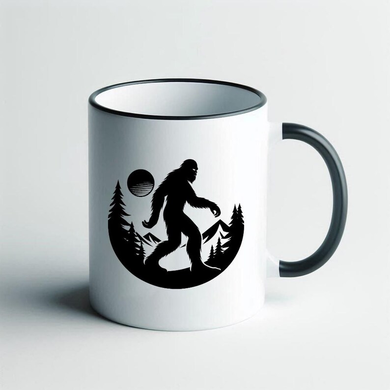 May include: White ceramic mug with a black rim and handle. The mug features a black silhouette of Bigfoot walking in front of a mountain and forest scene with a sun in the background.