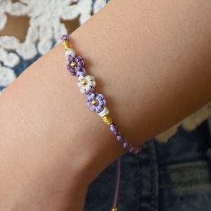 Peut inclure: Un bracelet macramé fait à la main avec trois breloques en forme de fleurs violettes, blanches et dorées. Le bracelet est réglable et semble être fait d'une matière tissée douce. Le bracelet est présenté sur le bras d'une personne.