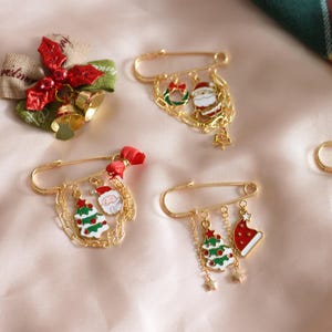 May include: A collection of gold-coloured Christmas brooches featuring festive charms such as Santa Claus, Christmas trees, wreaths, and bells. The brooches are arranged on a pale pink surface, with green plaid fabric in the background.