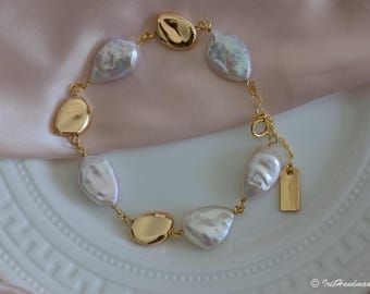 May include: A gold-toned bracelet featuring irregular, iridescent white pearls and gold-colored, organic-shaped links. The bracelet has a delicate chain and a small, rectangular gold tag. The jewelry is displayed on a white plate and a pink satin fabric.