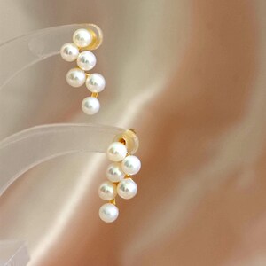 May include: Gold-coloured earrings with a cluster of white pearls. The earrings have a post-back fastening and a cascade design, with each earring featuring multiple pearls. The background is a soft, neutral colour.