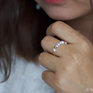 May include: A delicate gold chain ring with a row of small, white pearls. The ring is worn on a person's finger.
