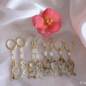 May include: Gold earrings featuring fish-shaped charms with an iridescent finish, complemented by beads and pearls. A pink flower rests above the earrings on a white satin backdrop. These earrings are a unique piece of handcrafted jewellery.
