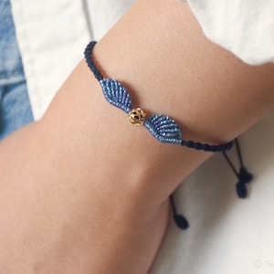 May include: A blue and black friendship bracelet with a gold bead in the center. The bracelet is made with a knotting technique and features two blue wings on either side of the bead.