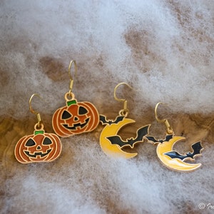 以下が含まれることがあります： ハロウィンをテーマにしたイヤリングのセットで、かぼちゃのデザインが2つ、三日月デザインが2つあります。かぼちゃはオレンジ色で、黒い目と大きな笑顔をしています。三日月は黄色で、黒いコウモリが周りを飛んでいます。