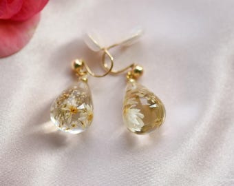 Puede incluir: Un par de pendientes dorados con colgantes en forma de lágrima. Cada colgante transparente contiene pequeñas flores blancas y detalles dorados. Los pendientes se muestran sobre una superficie de satén blanco.