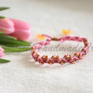 K&ouml;nnte beinhalten: Ein pinkfarbenes und braunes Freundschaftsarmband mit einem gewebten Muster. Das Armband ist mit einer Knotentechnik hergestellt.