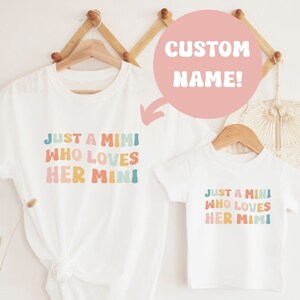 May include: Two white t-shirts with the text "Just a Mimi who loves her Mini" in rainbow colors. The text is in a playful, rounded font. The shirts are hanging on a wooden hanger. The larger shirt has the text "Custom Name!" written on it.