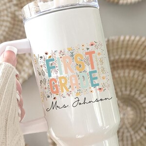 May include: White tumbler with a handle, featuring the words "FIRST GRADE" in colorful letters with floral accents. The name "Mrs. Johnson" is printed below. The tumbler has a clear lid and straw.