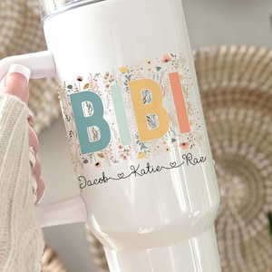 May include: White tumbler with a handle, featuring the name "BIBI" in colorful letters with floral accents. The text "DOUBLE SIDED PRINTING" is in a brown banner. The names "Jacob", "Katie", and "Rae" are also printed on the tumbler.