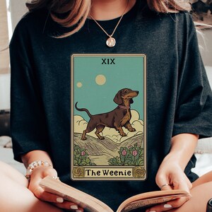 Pu&ograve; includere: Una maglietta nera con una grafica di un cane bassotto su una carta dei tarocchi. La carta &egrave; etichettata "XIX The Weenie".