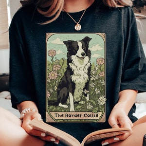 May include: A woman wearing a black t-shirt with a vintage-style illustration of a border collie dog. The illustration is surrounded by a light blue border and features the text "The Border Collie".