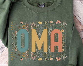 May include: A green sweatshirt with the word "OMA" in colorful letters, surrounded by floral designs. The sleeves are cuffed. The text "more colors available" is at the bottom.