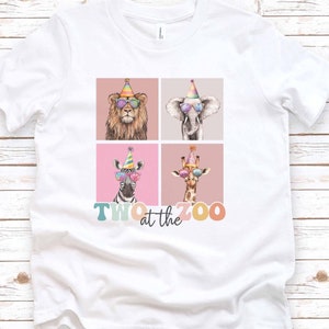 Op de afbeelding: Wit T-shirt met een afbeelding van vier dieren die feestmutsen en zonnebrillen dragen. De dieren zijn een leeuw, een olifant, een zebra en een giraffe. De tekst "TWO at the ZOO" staat onder de dieren.