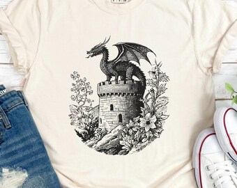 May include: Cream-colored t-shirt featuring a black and white illustration of a dragon perched atop a castle tower, surrounded by flowers. The shirt is paired with denim shorts and white sneakers with red accents, creating a casual, everyday look.
