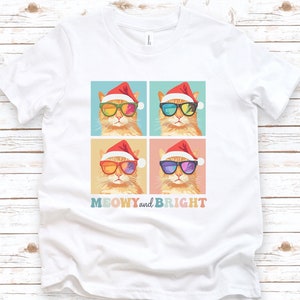 May include: White t-shirt featuring a colorful graphic of four cats wearing Santa hats and sunglasses. The cats have different colored lenses. The text "MEOWY and BRIGHT" is printed below the graphic. The shirt is displayed on a light wood background.