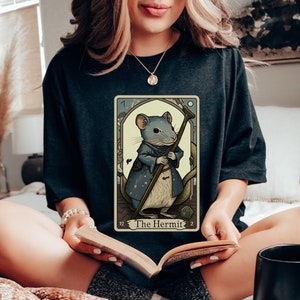 May include: A black t-shirt with a tarot card design featuring a mouse dressed as a hermit. The card is titled "The Hermit" and has a blue and gold color scheme.