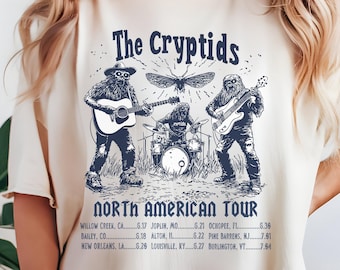 May include: A white t-shirt with a blue graphic print of a band called "The Cryptids" on a North American tour. The band features three members: a sasquatch playing a guitar, a mothman playing drums, and a bigfoot playing a bass guitar. The tour dates are listed below the band members.