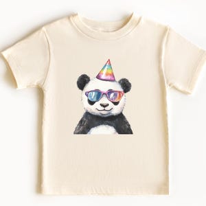 May include: Cream-colored t-shirt featuring a watercolor illustration of a panda wearing rainbow sunglasses and a party hat. The panda is black and white with a smiling expression. The shirt is short-sleeved and has a crew neck.
