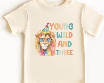 May include: A light beige t-shirt with a colorful lion wearing sunglasses and a party hat. The text "YOUNG WILD AND THREE" is printed on the shirt.