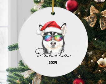 May include: White ceramic Christmas ornament featuring a husky dog wearing a Santa hat and rainbow sunglasses. The name "Dakota" is written in script, with the year "2024" below. The ornament hangs on a Christmas tree.