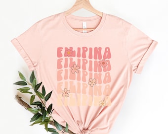 May include: A pink t-shirt with the word "FILIPINA" repeated in a retro font with flowers in between each word. The shirt is on a hanger against a white background.